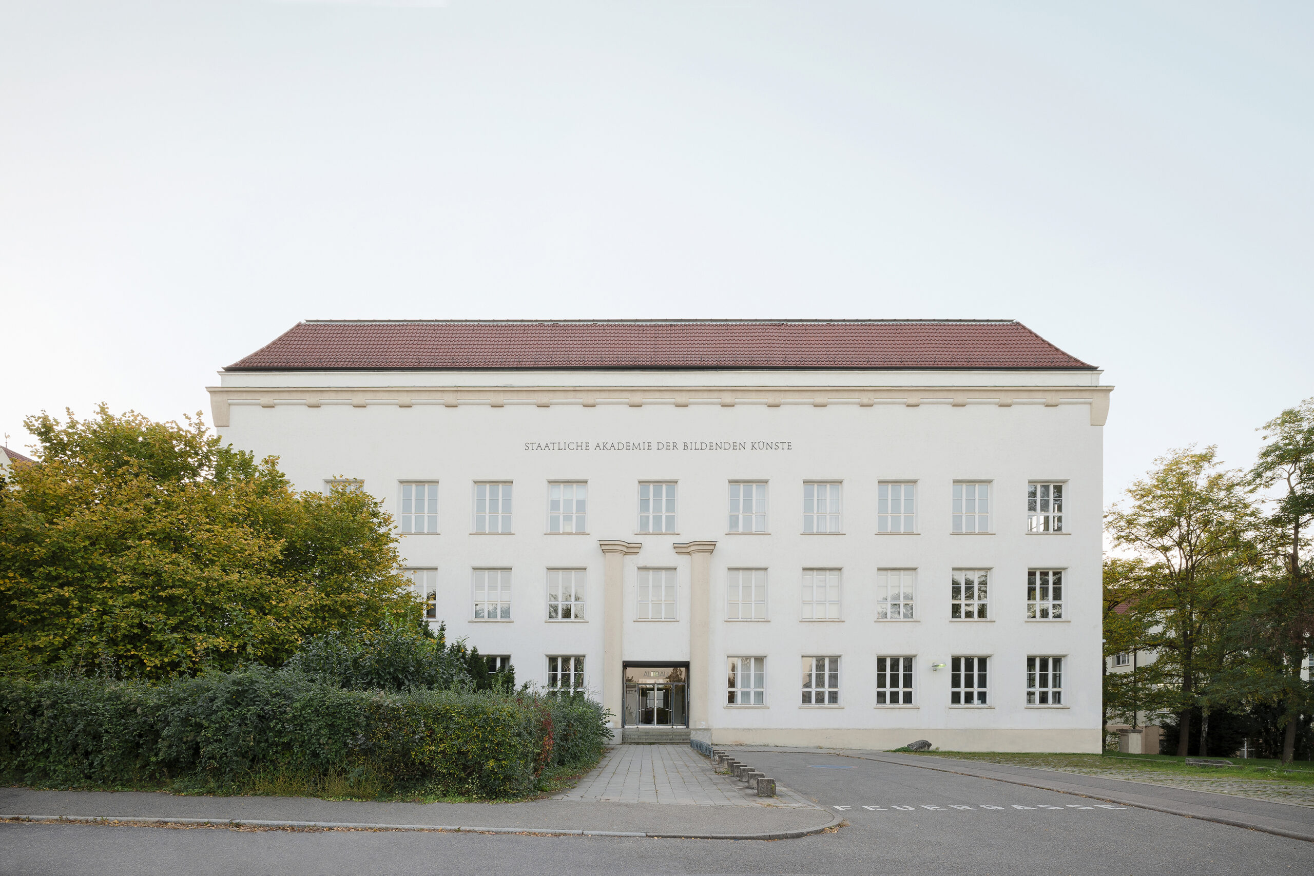 Ein symmetrisches weißes Gebäude mit einem roten Dach und der Aufschrift „Staatliche Akademie der Bildenden Künste“, umgeben von Grün und einem gepflasterten Gehweg, der zum Eingang führt.